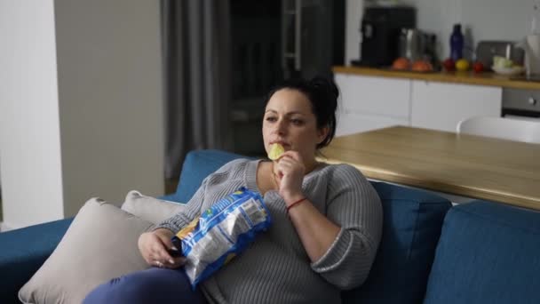Mujer con sobrepeso sentada en el sofá en casa, viendo la televisión y comiendo papas fritas — Vídeos de Stock