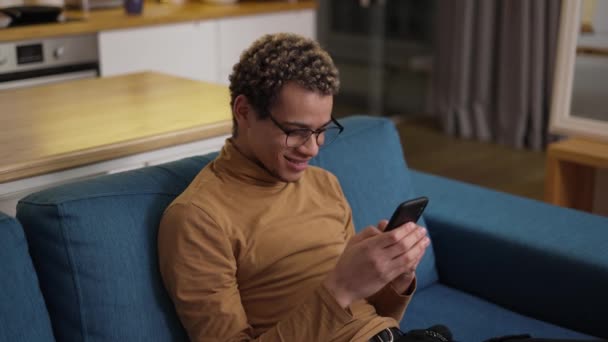 Krullende jongeman typen op zijn mobiele telefoon op de bank thuis — Stockvideo