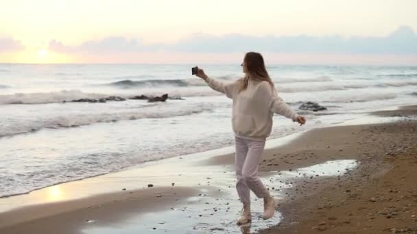Красивая молодая женщина крутится на берегу океана на закате, снимая себя на телефон — стоковое видео