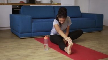 Oturma odasında paspasın üzerinde genç Afrikalı bir kadın, spor yapmak zor, esneme hareketleri yapıyor.