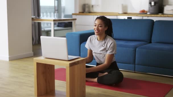 Leende afrikansk amerikansk kvinna pratar på videosamtal på laptop medan du sitter på yogamattan — Stockvideo