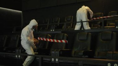 Koronavirüs pandemisi nedeniyle karantina. Korunma kostümü giymiş iki adam koltuklara uyarı kurdeleleri takıyor.