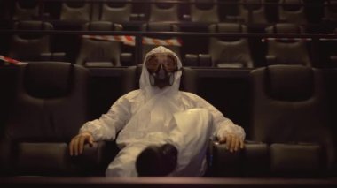Beyaz koruma kostümü ve solunum cihazıyla sinemada tek başına oturan bir adamın portresi.