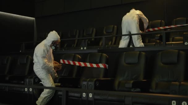 Quarentena devido a pandemia de coronavírus - dois homens em traje de proteção esticando fitas de advertência em assentos — Vídeo de Stock