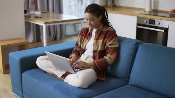 Mulher com dreadlocks está trabalhando em novo projeto e usando laptop no sofá na sala de apartamento — Vídeo de Stock