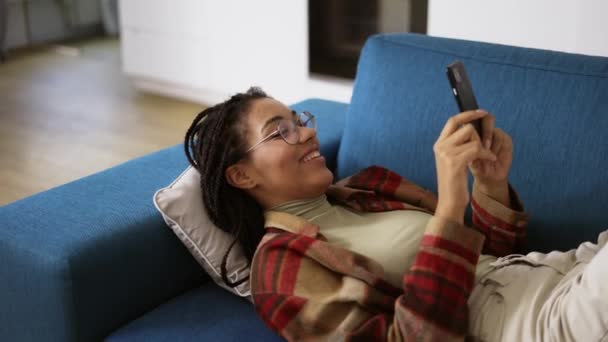Gelukkig Afrikaans Amerikaans meisje ontspannen op de bank thuis met smartphone — Stockvideo