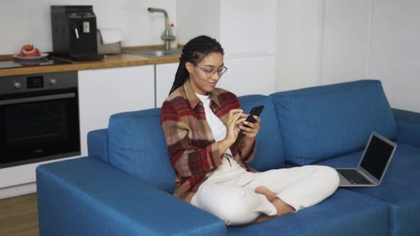 Fiatal nő használ okostelefon otthon egy kanapén laptop — Stock videók