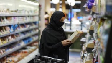Türbanlı ve koruyucu maskeli kadın süpermarkette market alışverişi yapıyor, mısır gevreğini seçiyor.