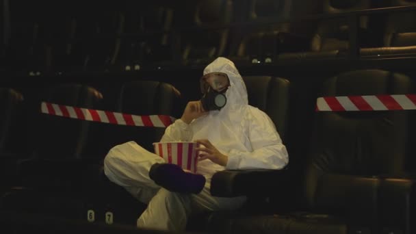 Beyaz koruma kostümü ve solunum cihazıyla sinemada tek başına oturmuş patlamış mısır yiyen bir adamın portresi. — Stok video