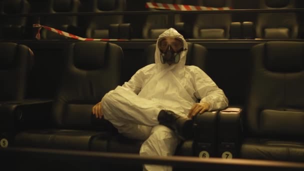 Uvolněný muž v bílém ochranném kostýmu a respirátoru sedí sám v kině — Stock video