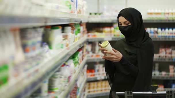 Mulher em hijab e máscara protetora fazendo compras, leva produto da prateleira em uma seção de leite — Vídeo de Stock