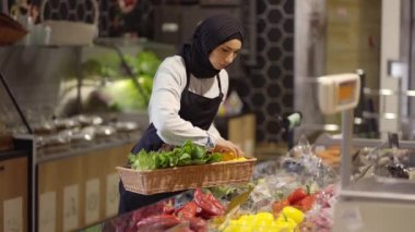 Önlüklü Müslüman kadın süpermarkette taze sebzeleri dolduruyor.