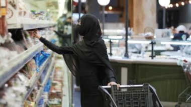 Tesettürlü ve koruyucu maskeli kadın alışveriş yapıyor, raftan ürün alıyor.