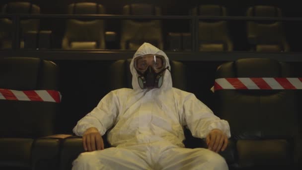 Portrait d'un homme en costume de protection blanc et respirateur assis seul au cinéma — Video