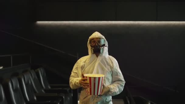 Mężczyzna w białym stroju ochronnym i respiratorze przyszedł sam do kina z wiadrem popcornu, w zwolnionym tempie. — Wideo stockowe