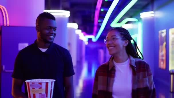 Χαρούμενη νεαρή Αφροαμερικάνα γυναίκα και άντρας που περπατούν δίπλα στο νέον Χολ με κουβά ποπ κορν, κουβεντιάζοντας στον κινηματογράφο — Αρχείο Βίντεο
