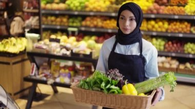 Başörtüsü içinde Müslüman bir kadın markette bir sepet taze sebzeyle yürüyor.