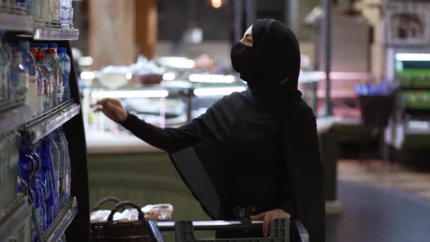 头戴头巾戴防护面具的女人去购物，从架子上拿一瓶水 — 图库视频影像
