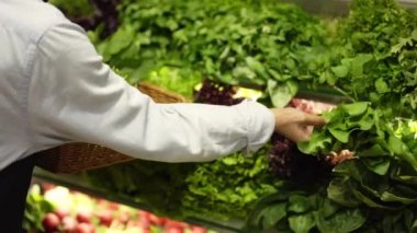 Önlüklü kadın süpermarketin rafındaki taze yeşillikleri dolduruyor.