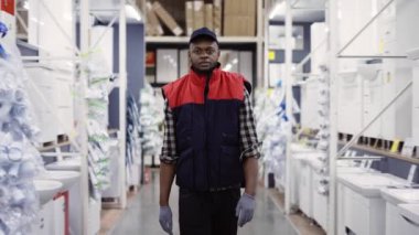 Satıcı hırdavatçıda sıraların arasında yürüyor, kameraya bakıyor.