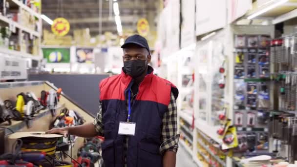 Продавець в масці стоїть між рядками в магазині обладнання, дивлячись на камеру — стокове відео