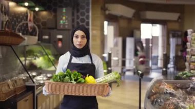 Türbanlı Müslüman kadın markette bir sepet taze sebzeyle yürüyor.