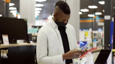 Maskeli Afrikalı Amerikalı bir adam dükkandan yeni bir cep telefonu seçiyor, nasıl çalıştığını kontrol ediyor.