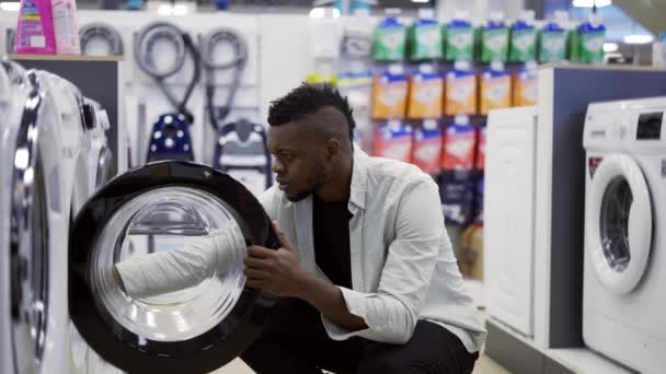 El hombre afroamericano está eligiendo la lavadora en la tienda de electrodomésticos, viendo la muestra de la exposición — Vídeos de Stock