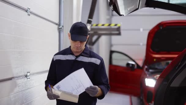 Hombre de uniforme en un taller de reparación de automóviles caminando por ahí, mirando a sus notas — Vídeo de stock