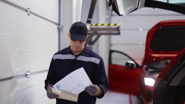 Hombre de uniforme en un taller de reparación de automóviles caminando por ahí, mirando a sus notas — Vídeos de Stock