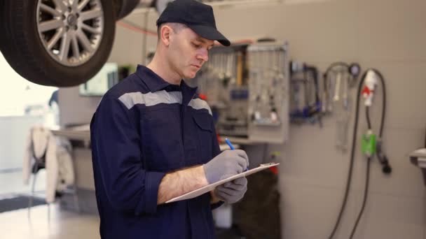 Mecánico en un taller de reparación de automóviles haciendo notas a su tableta — Vídeos de Stock