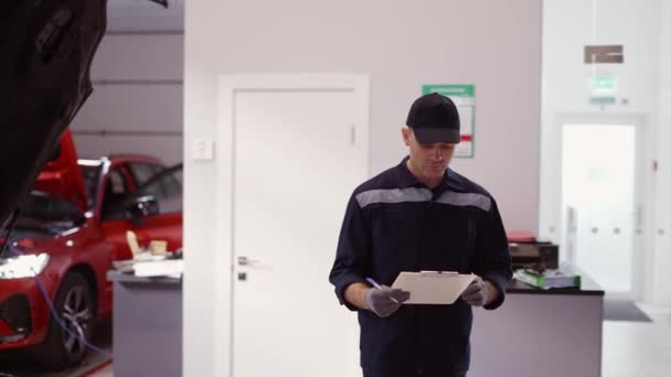 Hombre de uniforme en un taller de reparación de automóviles caminando por ahí, tomando notas en su tableta — Vídeos de Stock