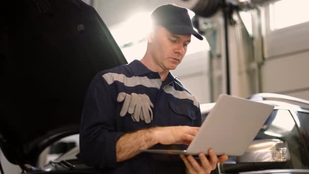 Mécanicien de voiture travaillant sur ordinateur portable dans le service de réparation automobile, pencher sur une voiture, vue à faible angle — Video