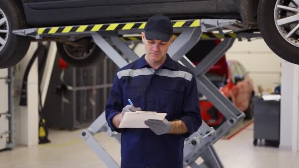 Un mecánico de coche de pie junto al coche levantado, tomando notas en una tableta — Vídeos de Stock