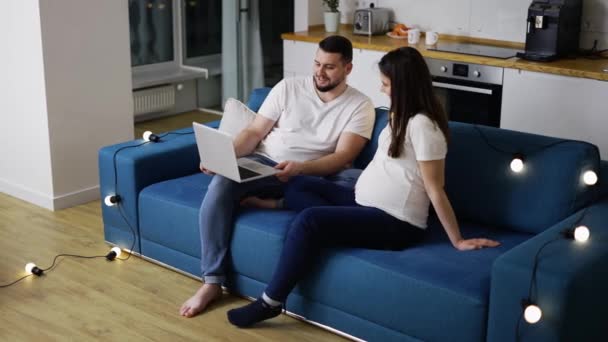 Υπέροχο έγκυο ζευγάρι χρησιμοποιώντας ένα φορητό υπολογιστή κάθεται στον καναπέ σε ένα νέο σαλόνι με γιρλάντες — Αρχείο Βίντεο