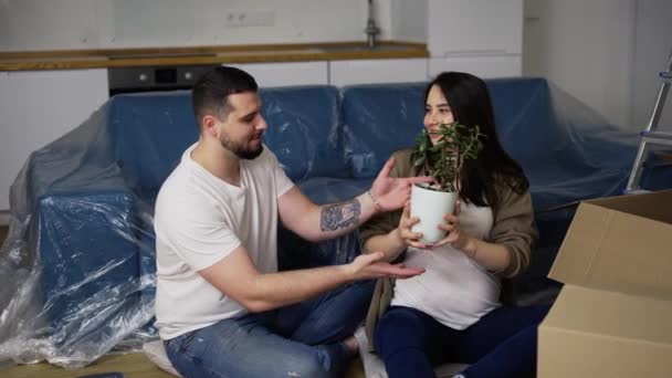 Lächelndes Paar sitzt im Wohnzimmer auf dem Boden und packt Pflanze aus der Schachtel — Stockvideo