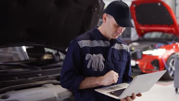 Mecánico de coche que trabaja en el ordenador portátil en el servicio de reparación de automóviles, se apoyan en un coche — Vídeos de Stock