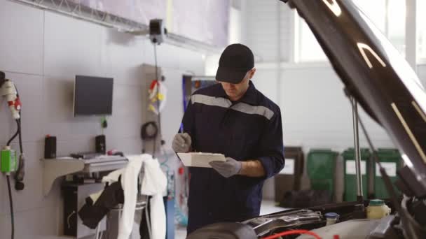 Mécanicien dans un atelier de réparation automobile vérifiant le moteur en prenant des notes sur sa tablette — Video