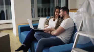 Güzel hamile bir kadın ve pozitif bir adam yeni oturma odasındaki koltukta dizüstü bilgisayar kullanıyorlar.