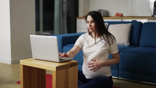 Una donna incinta in abiti da casa è seduta sul pavimento e utilizza un computer portatile — Video Stock