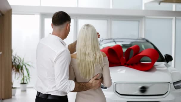 Um homem faz surpresa para uma mulher loira, fecha os olhos das mulheres pelas costas carro novo com arco vermelho, visão traseira — Vídeo de Stock