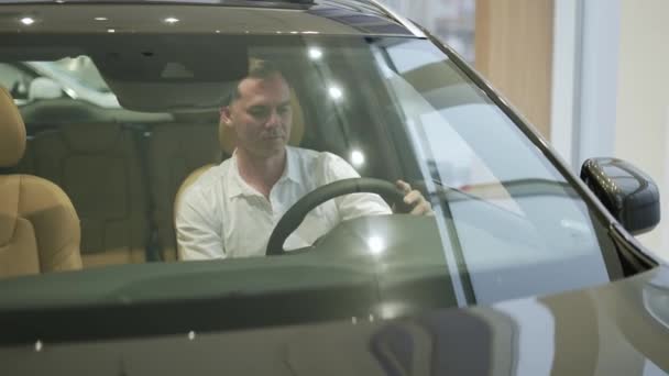 Cliente na camisa branca sente-se no salão do carro, quer comprar um novo automóvel no showroom — Vídeo de Stock