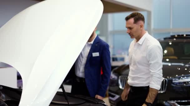 Vendedor profesional que muestra el motor del coche bajo la capucha abierta a la pareja en la sala de exposición automática — Vídeos de Stock