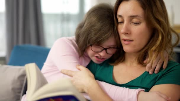 Ung mor läser en bok för sin dotter med ner syndrom medan de sitter på soffan i rummet — Stockvideo