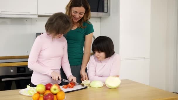 Dvě dívky s Downovým syndromem krájení zeleniny s jejich matkou v kuchyni — Stock video