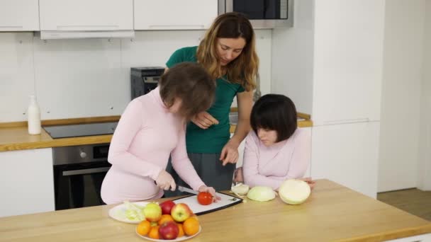 Δύο κορίτσια με σύνδρομο Down κόβουν λαχανικά με τη μητέρα τους στην κουζίνα στο σπίτι — Αρχείο Βίντεο