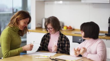 Down sendromlu iki kız mutfakta öğretmen ya da annenin yanında resim yapıyor, ağır çekimde.