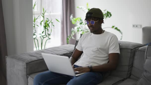 魅力的なアフリカ系アメリカ人男性で眼鏡でザソファに座って使用してノートパソコンで自宅 — ストック動画