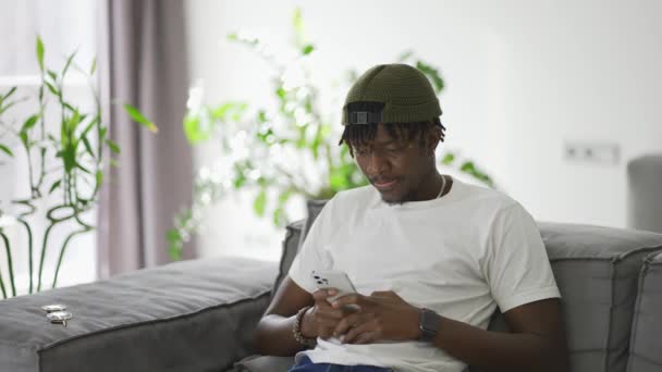 アフリカ系アメリカ人の男性がリビングでソファに座っている間にスマートフォンを使って — ストック動画