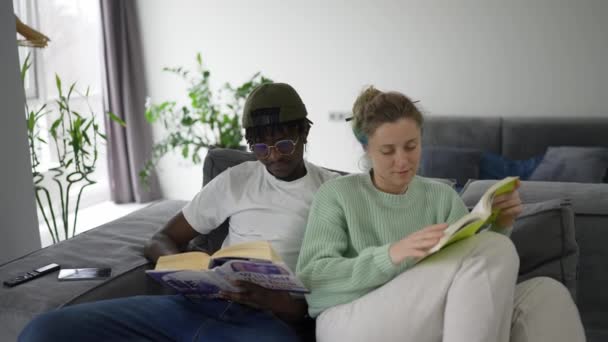 Νεαρό Μικτό Ζευγάρι Κάθεται Στον Καναπέ Τους Και Διαβάζει Βιβλία — Αρχείο Βίντεο